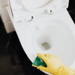 From above crop unrecognizable cleaner in rubber yellow gloves using sponge to wipe off toilet bowl in restroom