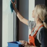 A woman in overalls cleans a window with a cloth in natural light.